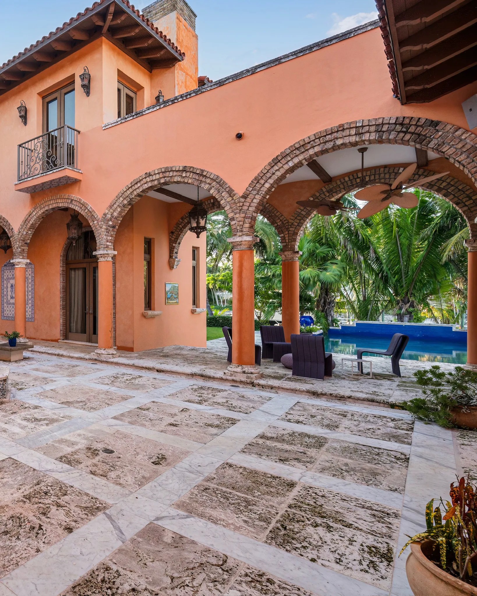 Terracotta Mediterranean home with brick arches framing a tiled patio, outdoor seating, and a pool amid tropical palms.