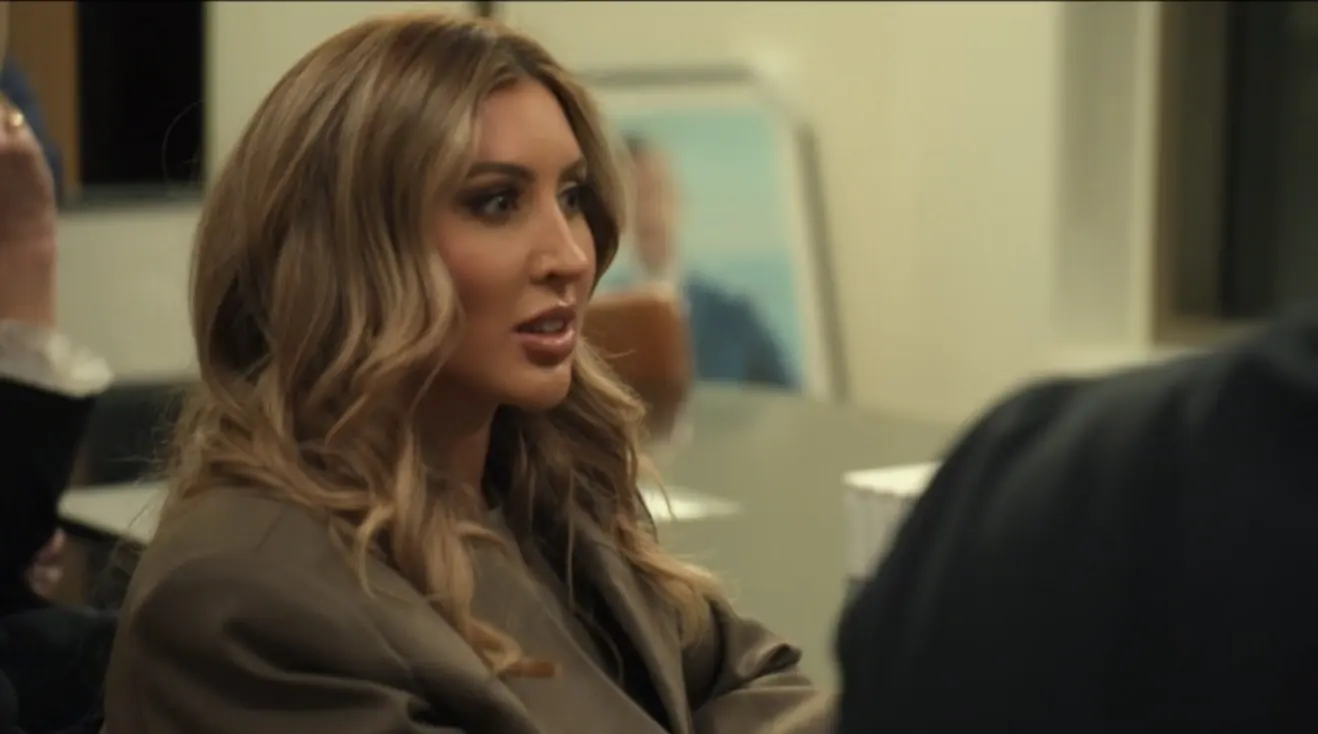 Woman in a taupe blazer speaking indoors, with long wavy blonde hair.
