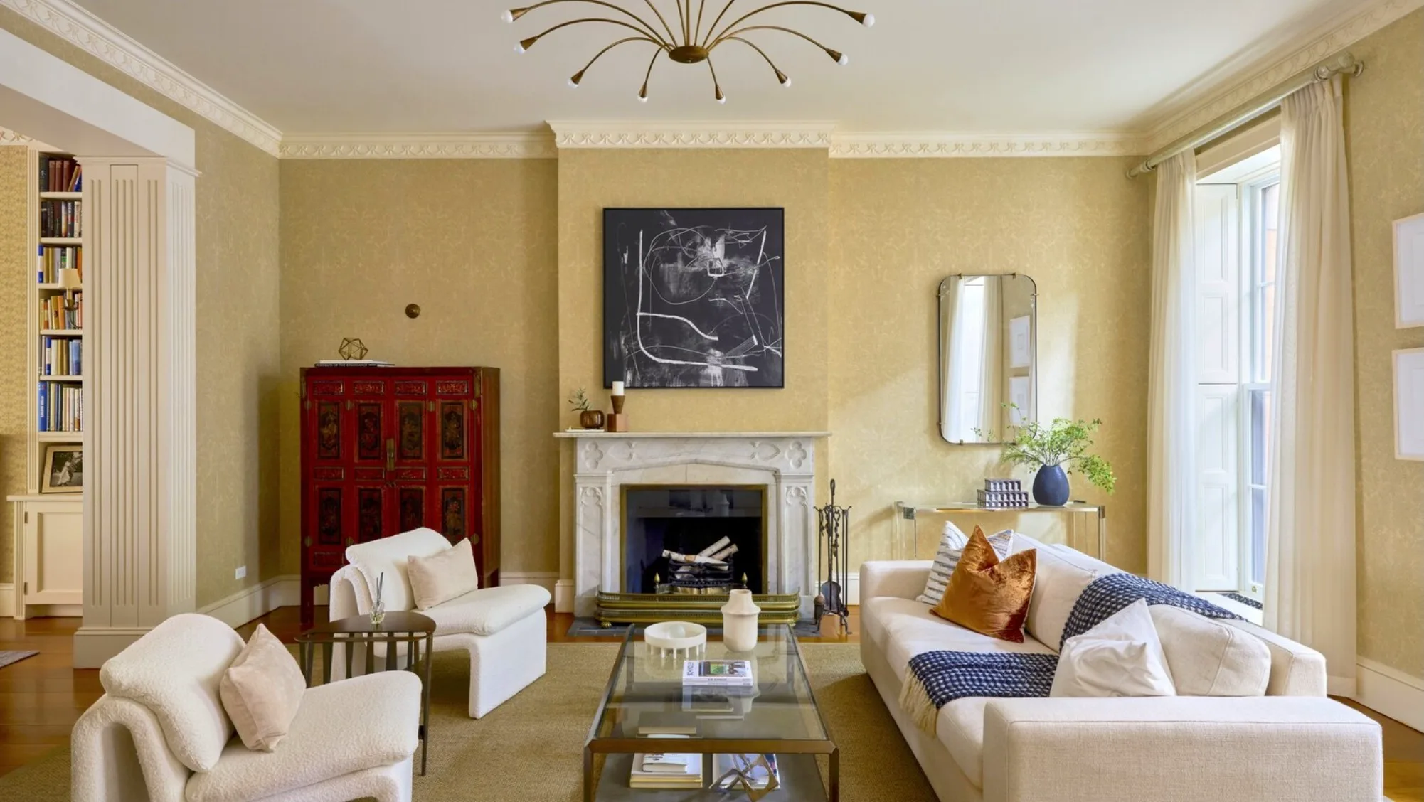 Elegant living room with a carved white fireplace, red antique cabinet, and tall sunlit windows.