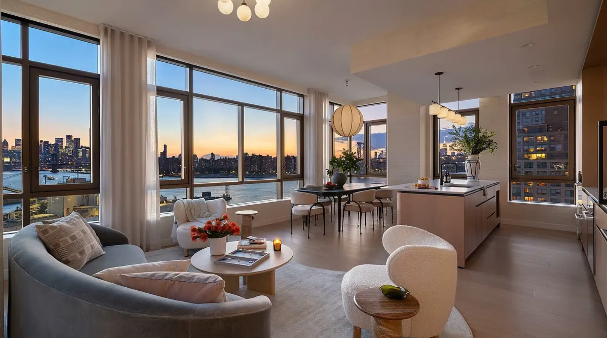 Open-plan apartment living room and kitchen with floor-to-ceiling windows overlooking the river and skyline at dusk.