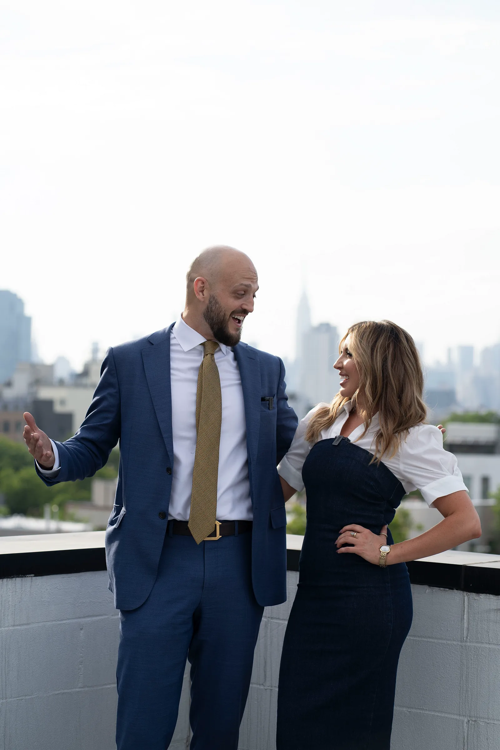Two people chat on a rooftop, with a city skyline behind them.