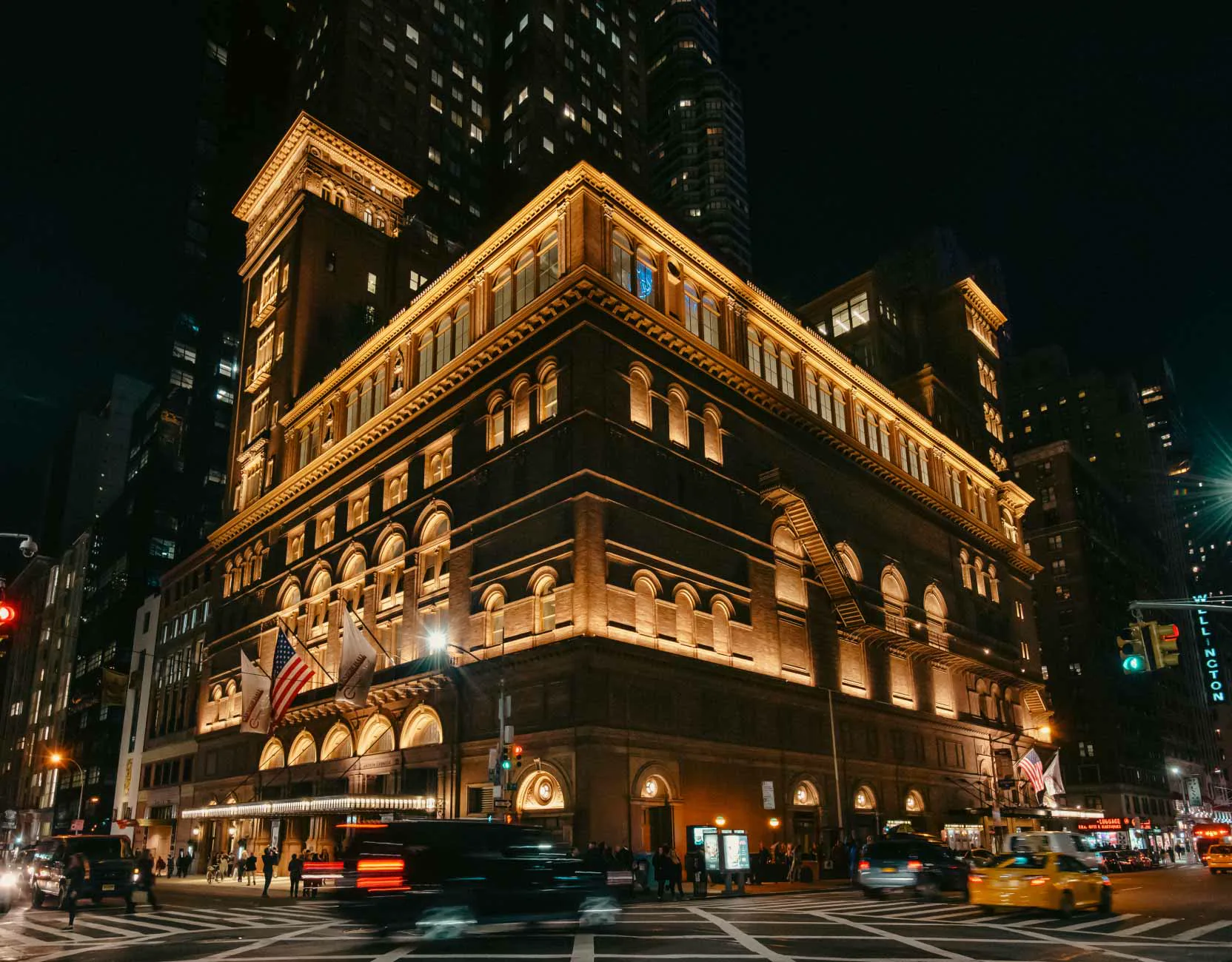 Steinway Hall’s ornate, illuminated corner facade at night, with the modern Steinway Tower behind it.