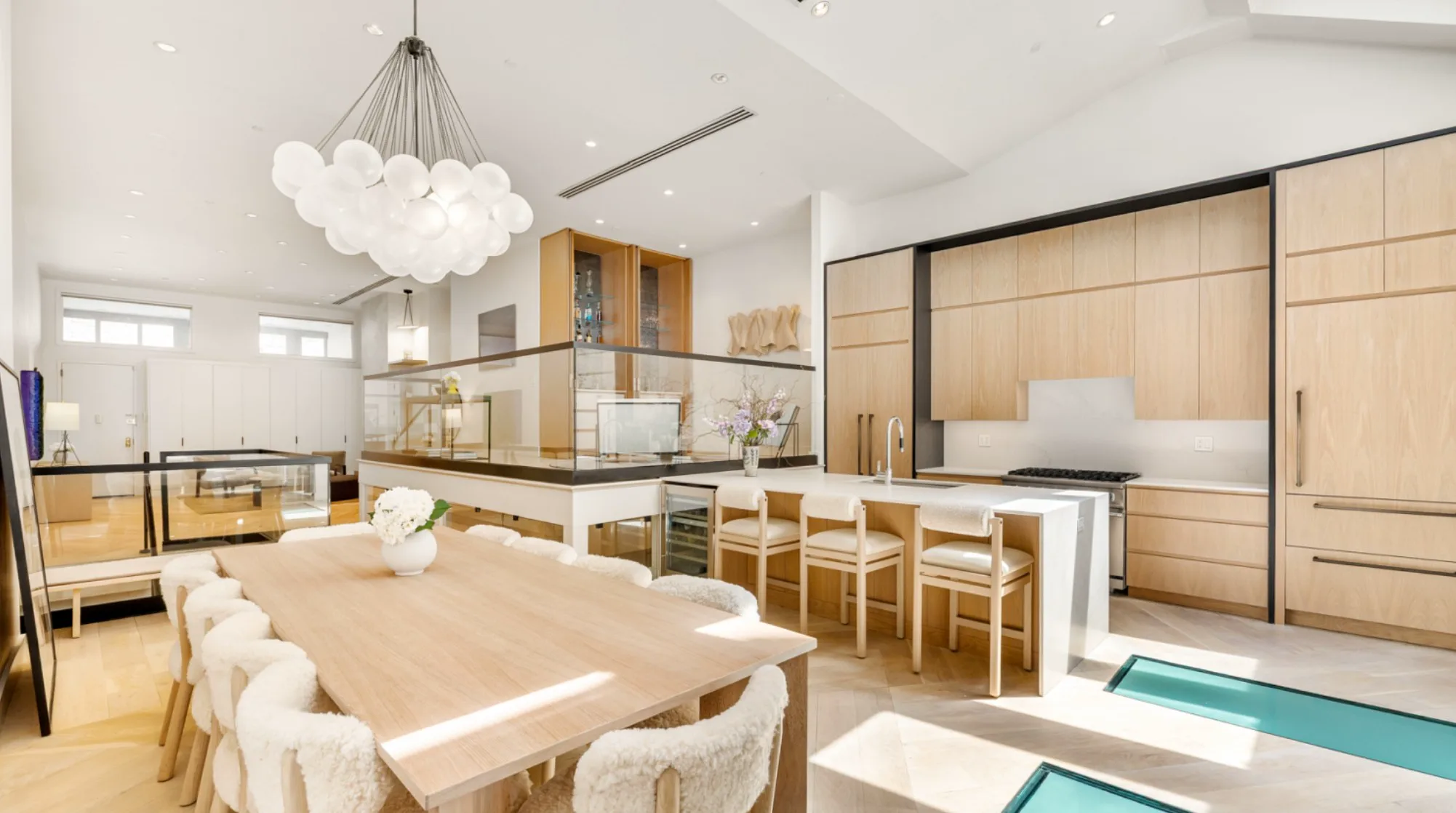 Open-plan kitchen and dining area with pale wood cabinetry, glass partitions, and turquoise water visible at floor level.