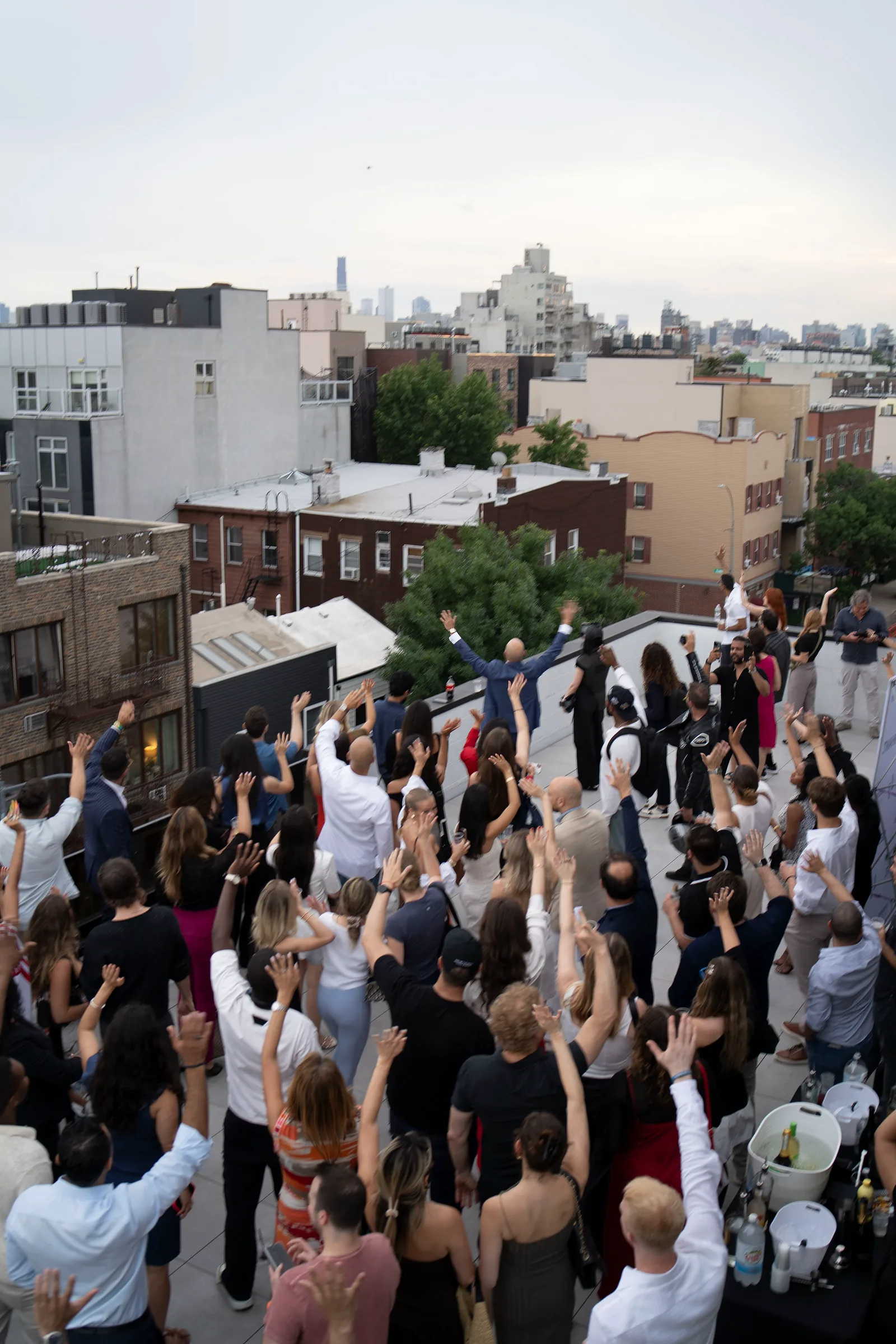 Crowd celebrates at a rooftop launch event overlooking Brooklyn rooftops and the city skyline.