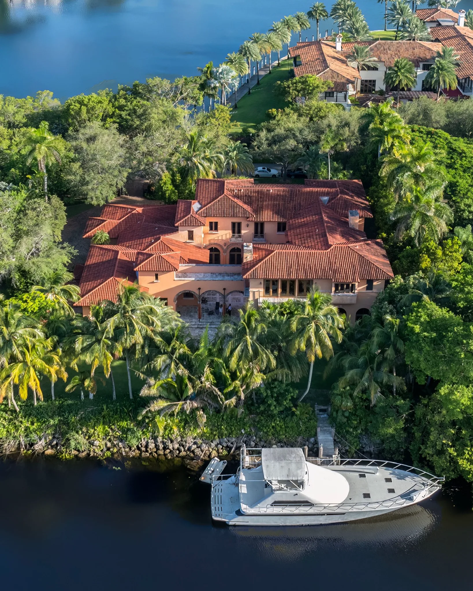 Aerial view of a terracotta-roofed waterfront mansion surrounded by palms, with a white yacht moored nearby.