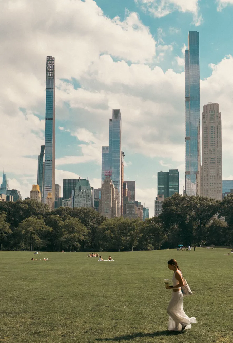 Central Park lawn with Midtown Manhattan skyscrapers rising behind the trees.
