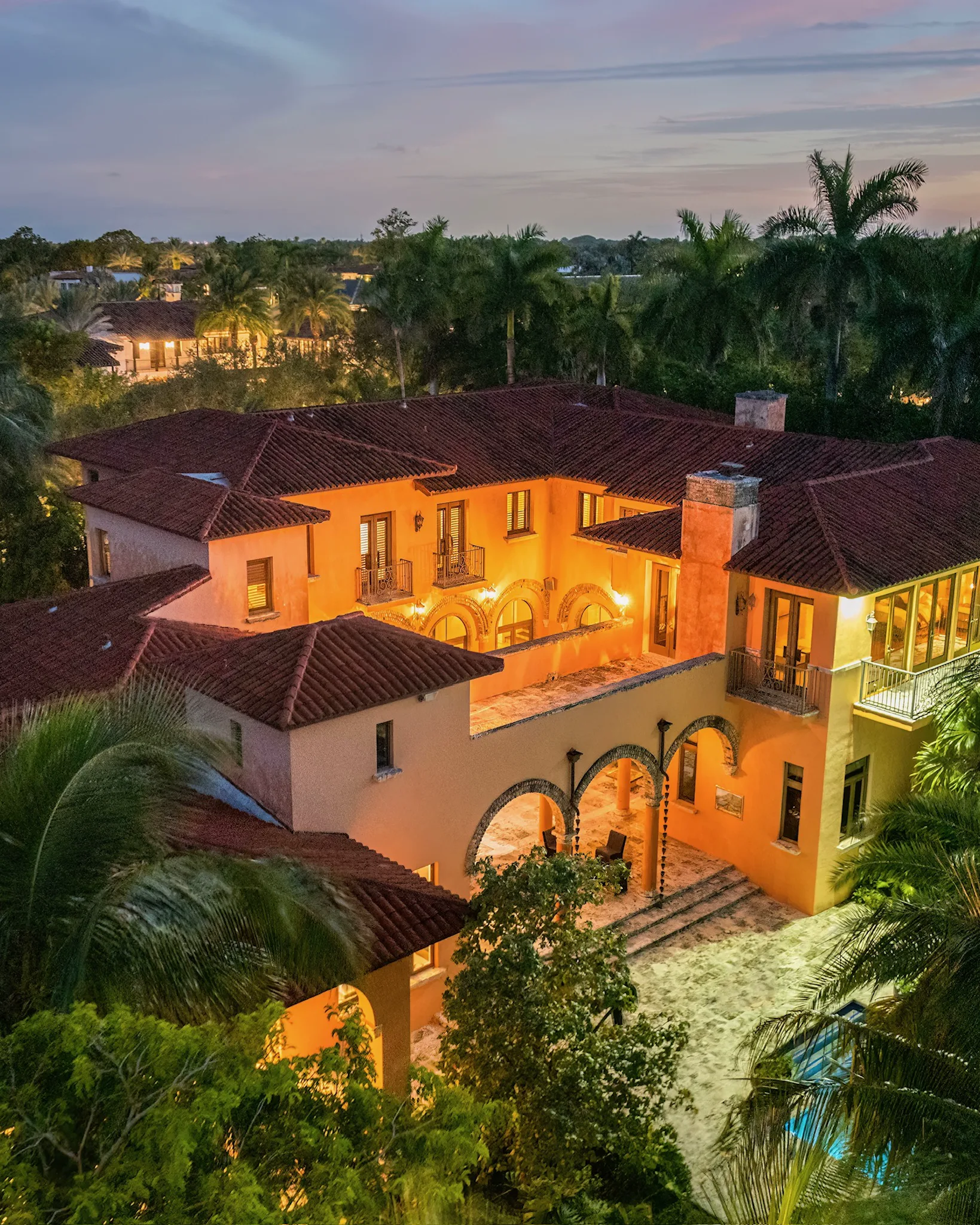 Villa De Casuarina at dusk, with illuminated arches, red tile roofs, palms, and a pool.