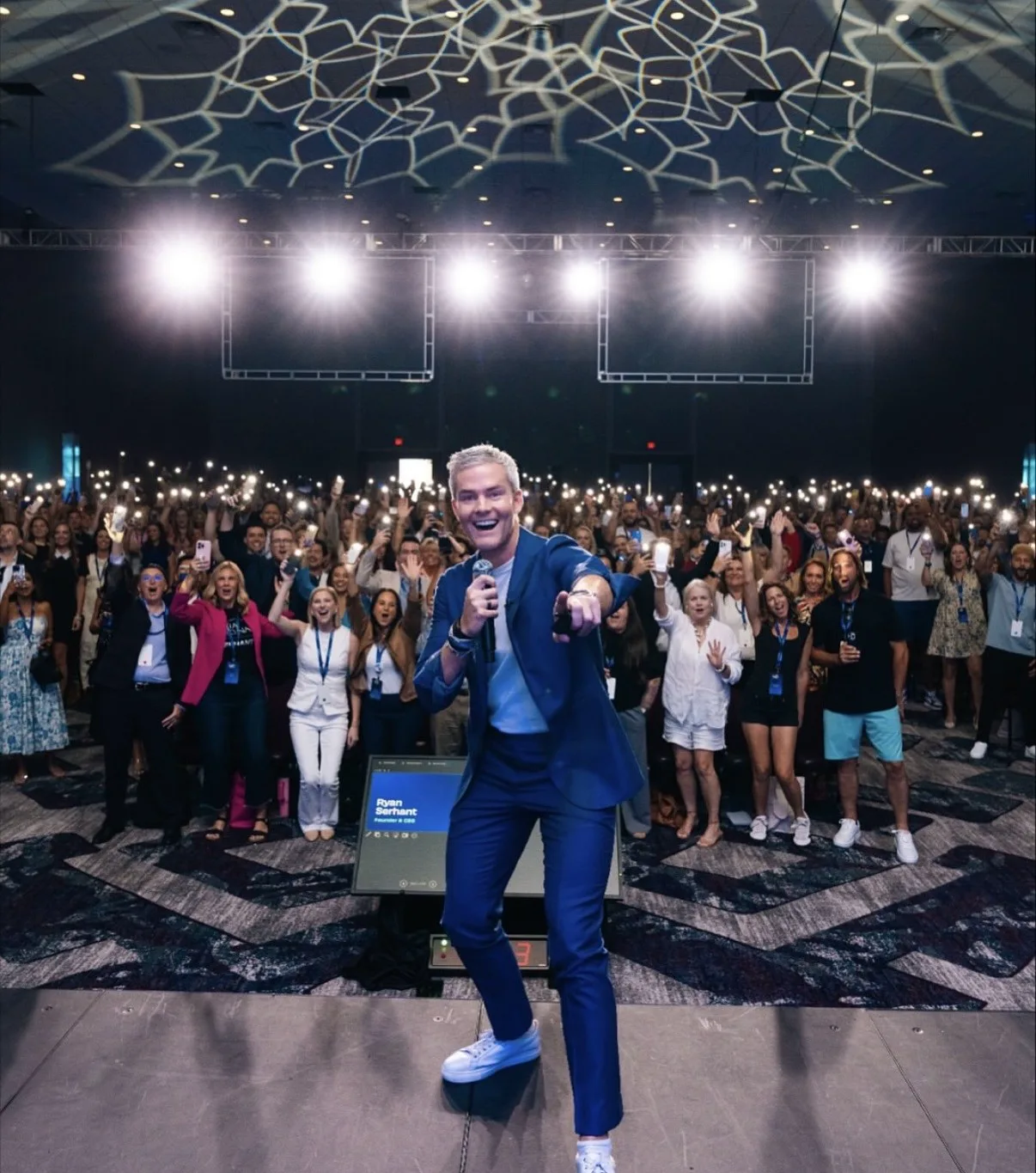 Ryan Serhant addresses a cheering crowd at a brightly lit real estate conference.