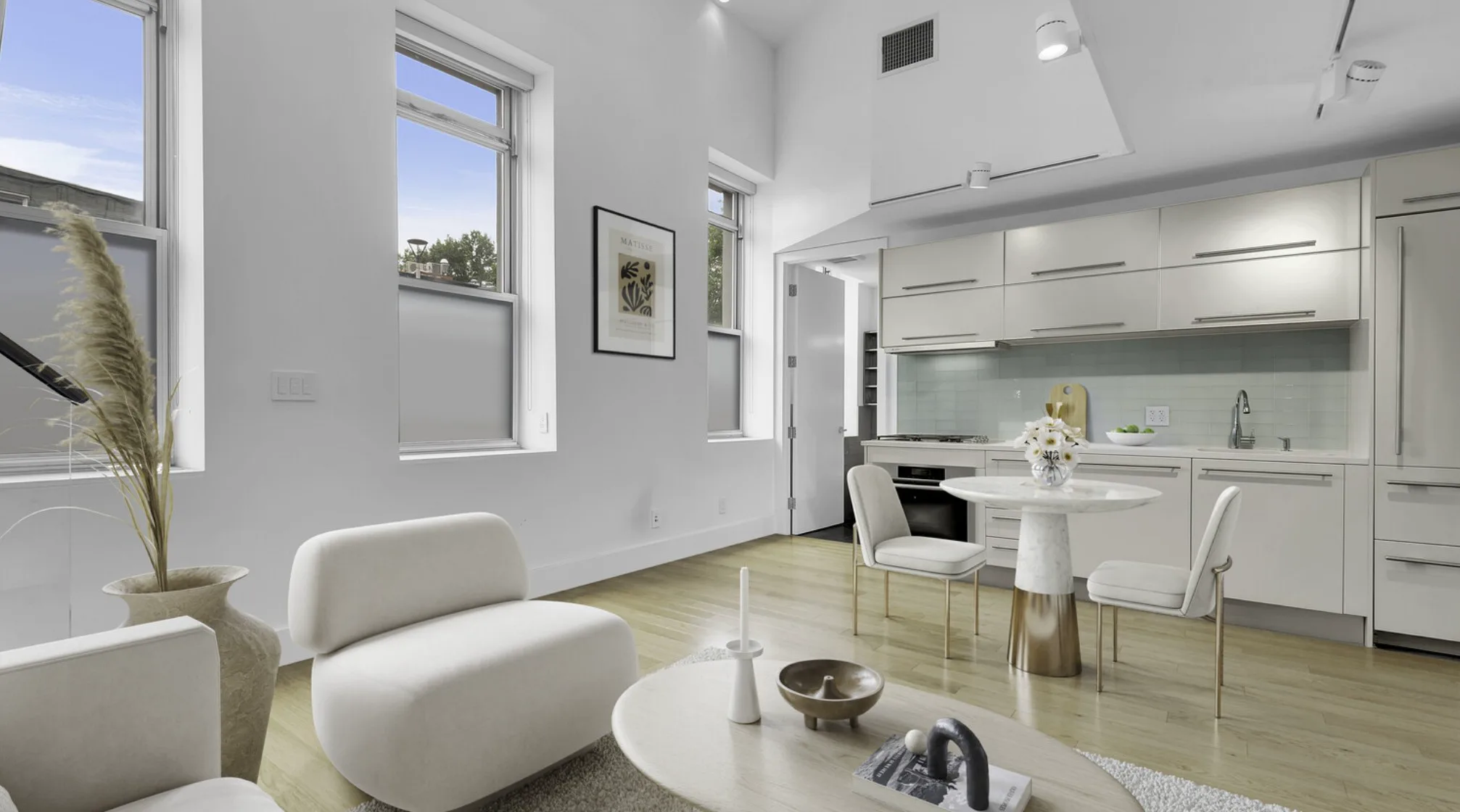 Modern white kitchen and living area with large windows, a round dining table, and sculptural chairs.