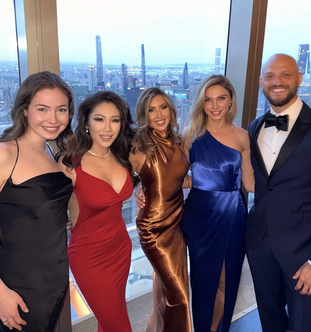 Five adults in formal attire pose before a floor-to-ceiling window overlooking the New York City skyline.